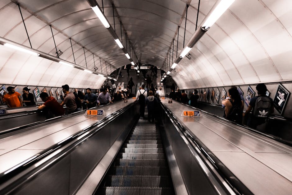 People standing and walking on the platform of a underground train station, preparing to board or disembark from the trains on either side. Some individuals are carrying luggage or backpacks, while ot