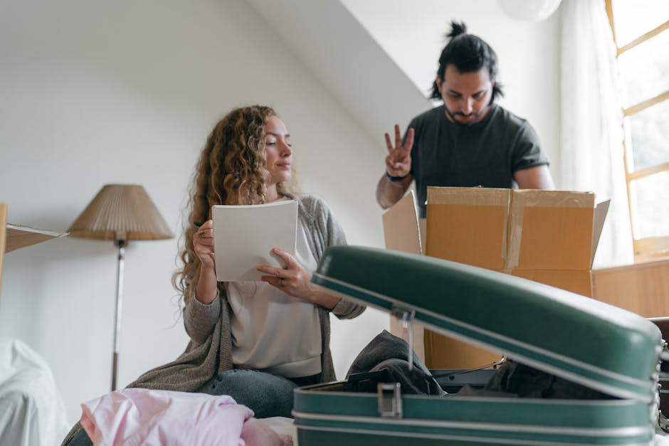 A woman with long curly hair, dressed in a light-colored top and grey cardigan, holds an open cardboard box while sitting on a bed in a well-lit room with natural daylight streaming through a window.